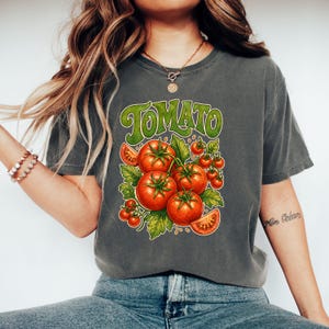 May include: A charcoal gray t-shirt featuring a graphic print of red tomatoes on green vines with the word TOMATO in large green letters at the top. The design includes several whole tomatoes and two sliced tomato segments with green leaves.