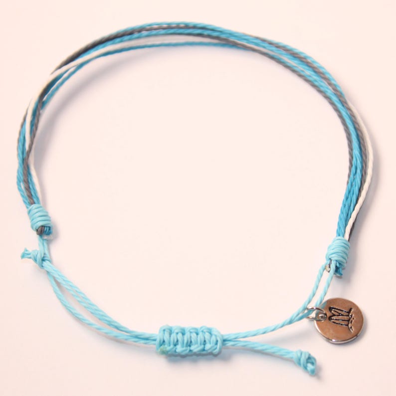 May include: A light blue, white, and gray braided bracelet with a silver charm. The charm has the letter "M" on it.