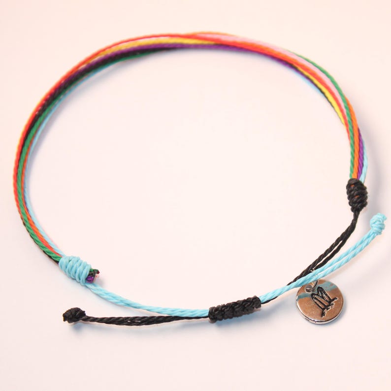 May include: A colorful braided bracelet with a silver charm featuring the letter "M". The bracelet is made of multiple strands of colorful thread, including red, orange, yellow, green, blue, and purple.