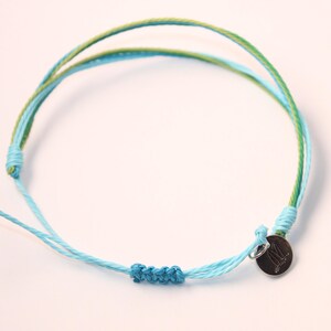 May include: A blue and green friendship bracelet with a silver charm. The charm has a small design on it.