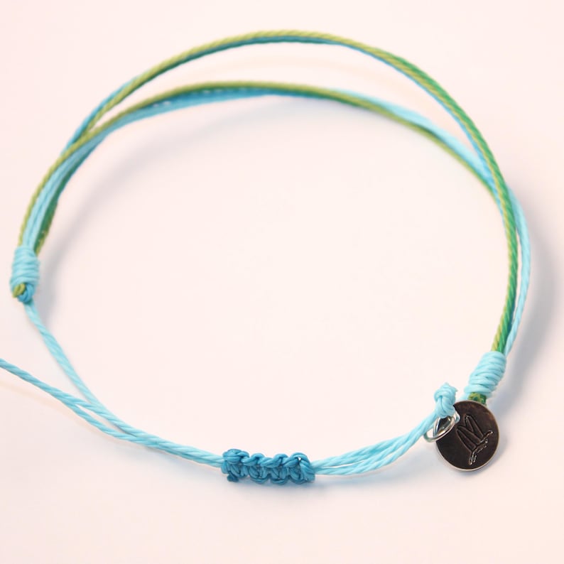 May include: A blue and green friendship bracelet with a silver charm. The charm has a small design on it.
