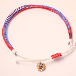 May include: A white, red, and blue braided bracelet with a silver charm. The charm has a design of two birds facing each other.