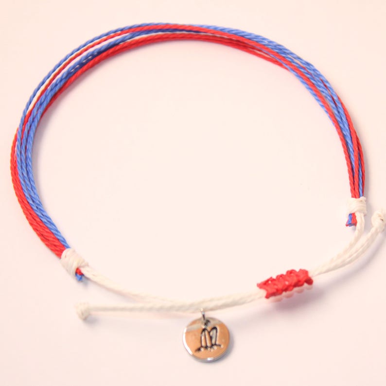 May include: A white, red, and blue braided bracelet with a silver charm. The charm has a design of two birds facing each other.