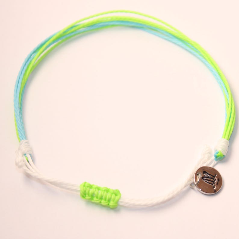 May include: A white, green, and blue friendship bracelet with a silver charm. The charm has a symbol on it.