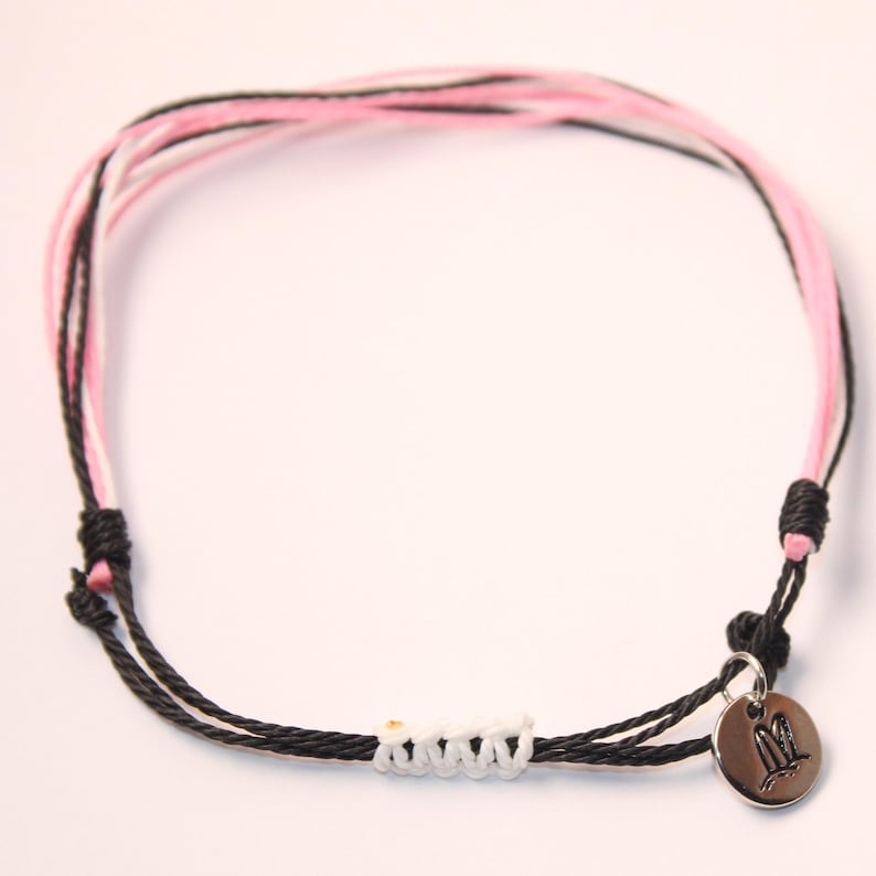 May include: A black and pink friendship bracelet with a silver charm featuring a bird design. The bracelet is made of braided cord and has a white knot detail.
