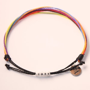 May include: A colorful rainbow string bracelet with a black cord and white beads. The bracelet has a silver charm with the astrological symbol for Gemini.