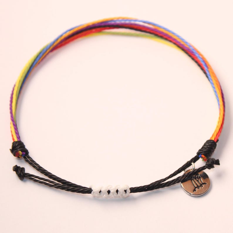 May include: A colorful rainbow string bracelet with a black cord and white beads. The bracelet has a silver charm with the astrological symbol for Gemini.