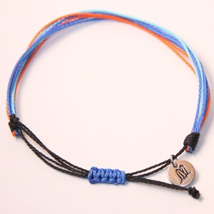 May include: A blue, orange, and light blue woven friendship bracelet with a black cord and a silver charm with a double surfboard design.