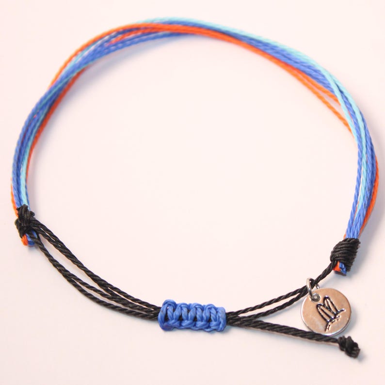 May include: A blue, orange, and light blue woven friendship bracelet with a black cord and a silver charm with a double surfboard design.