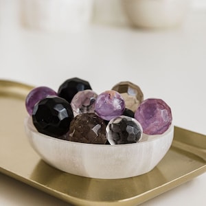 May include: A white selenite bowl filled with various gemstone spheres, including amethyst, clear quartz, smoky quartz, and black obsidian.