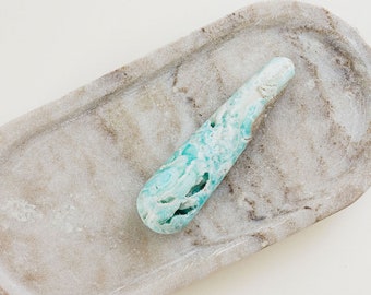 May include: A light blue and white crystal gemstone in the shape of a teardrop, resting on a light gray marble tray.