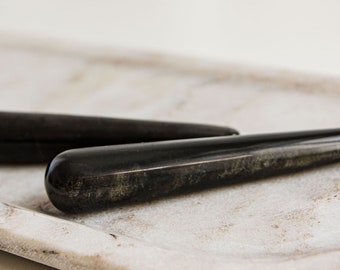 May include: Two black smooth stones with a slight shimmer, shaped like long, thin cylinders, are resting on a white ceramic tray.