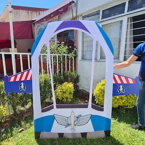 Può includere: Un ritaglio di cartone blu e bianco di un'astronave con una finestra viola. L'astronave ha un'ala a strisce rosso, bianco e blu su ogni lato. L'astronave ha un logo con un razzo e ali sul davanti.