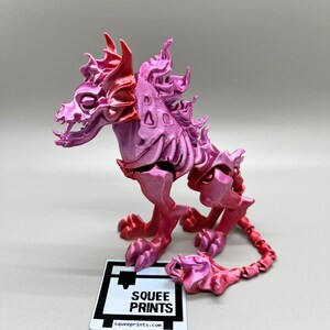 May include: A pink and red 3D printed articulated skeletal creature with a long tail. The creature has a skull-like head with horns and a fiery mane. The creature is posed in a standing position with its tail curled around its legs. The creature is printed by "Squee Prints".