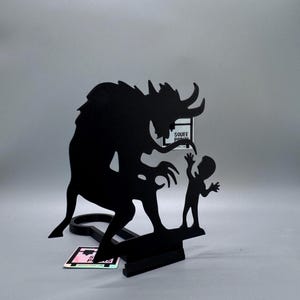 May include: Black metal sculpture depicting a horned creature reaching toward a small figure with outstretched arms. The sculpture is mounted on a black base with the text "SOUEE PRINTS" visible. The overall design has a fantasy theme.