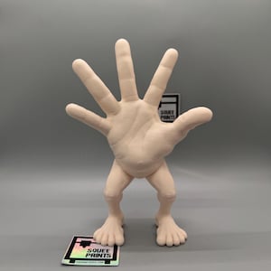 May include: A 3D printed figurine of a hand with five fingers standing on two legs. The figurine is made of a light beige plastic and is about 10 inches tall.