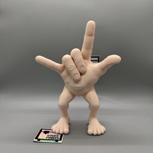 May include: A 3D printed figurine of a hand making the 'I love you' sign with its fingers. The hand is attached to a small body with legs and feet. The figurine is made of a light pink plastic.