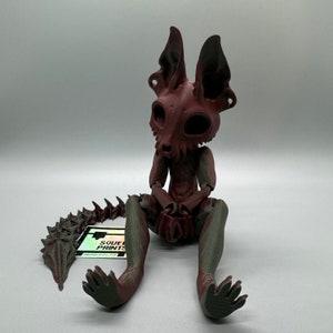 May include: A 3D printed figurine of a creature with a skeletal head, large ears, and a long, segmented tail. The creature is sitting with its legs crossed and its hands clasped in its lap. The figurine is painted in shades of brown and green.