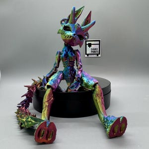 May include: A multi-colored iridescent figurine of a sitting creature with a spiky tail. The creature has a skeletal appearance with horns and a pointed head. The figurine is sitting on a black circular base. The text "SOUFFÉ PRINTS" is visible on a sticker.