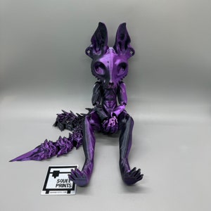 May include: A purple and black 3D printed figurine of a creature with a long tail, large ears, and a skeletal appearance. The figurine is sitting with its legs crossed and its hands clasped together. The figurine is made by "Squee Prints".