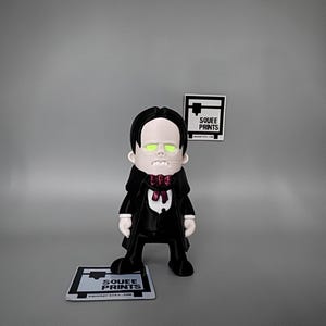 May include: A 3D-printed figurine of a vampire character, dressed in a black suit with a red bow tie and cape. The figurine has glowing green eyes and is standing on a small platform with the text "Squee Prints".