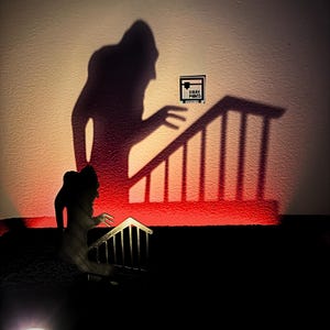May include: A silhouette of a figure climbing a staircase, with a red glow behind it. The figure is holding a small sign that says "SOUFE PRINT".