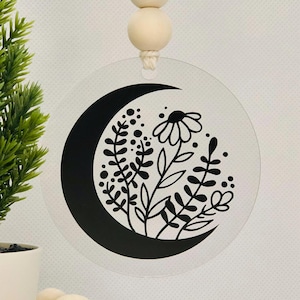 Puede incluir: Un adorno de acrílico transparente con una luna creciente negra y un diseño floral. El adorno cuelga de una cuerda con tres cuentas de madera.