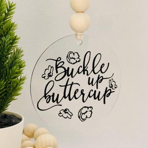 May include: A clear round acrylic ornament with the text "Buckle up buttercup" in black script. The ornament is hanging from a string with three wooden beads.