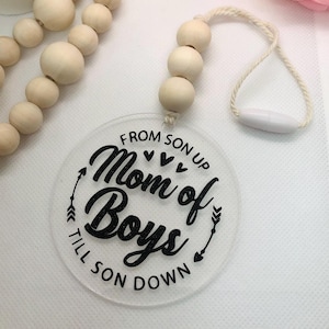May include: A clear, circular car charm with the text "Mom of Boys" in black script, surrounded by arrows and hearts. The charm hangs from a string of wooden beads and a white clasp.