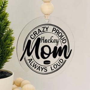 May include: A clear acrylic bauble with a black circle design that says "Crazy Proud Hockey Mom Always Loud". The bauble is hanging from a string with three wooden beads.