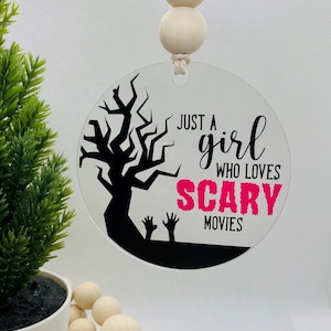 May include: A round, clear acrylic bauble with a black silhouette of a tree and the text "Just a girl who loves scary movies" in pink and black lettering.