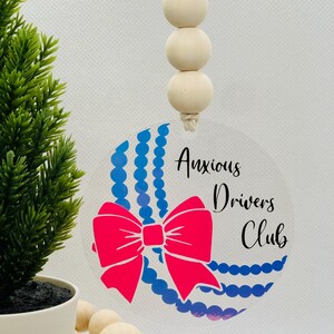 May include: A clear, circular car charm with the words "Anxious Drivers Club" in black script. It features a bright pink bow and blue beaded accents. The charm hangs from a string with wooden beads.