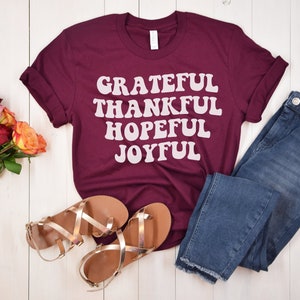 May include: Maroon t-shirt with the text "GRATEFUL THANKFUL HOPEFUL JOYFUL" in white. The shirt is laid flat on a white wooden surface with a pair of gold sandals, blue jeans, and a bouquet of roses.