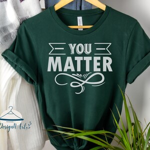 May include: A dark green t-shirt with the text "You Matter" in white lettering. The text is framed by a white arrow and a decorative swirl.