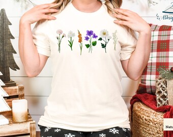 Op de afbeelding: Crèmekleurig T-shirt met een rij kleurrijke bloemenillustraties. De bloemen omvatten witte, roze, oranje en paarse bloemen met groene stelen. Het shirt is gemaakt van een zacht materiaal en is geschikt voor vrijetijdskleding.