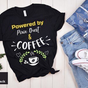 Może przedstawiać: Czarny T-shirt z napisem "Powered by Pixie Dust & COFFEE" w żółtych i białych literach. Koszulka ma zielony wzór liści i grafikę filiżanki kawy.