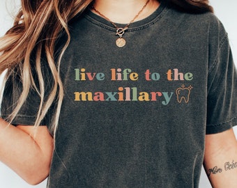 Op de afbeelding: Een donkergrijs T-shirt met de tekst "live life to the maxillary" in regenboogkleuren en een tandgrafiek.