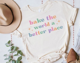 May include: A beige t-shirt with the text "bake the world a better place" in rainbow colors. The text is in a retro font style. The shirt is laid flat on a white surface with a brown hat, green plant, and brown shoes.