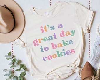 May include: A white t-shirt with the text "it's a great day to bake cookies" in rainbow colors. The shirt is laid flat on a white surface with a brown hat, a pair of brown shoes, and a pair of blue jeans with a white beaded necklace.