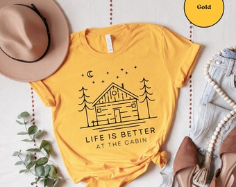 May include: A yellow t-shirt with a black outline of a cabin, trees, and a crescent moon. The text "Life is better at the cabin" is printed below the image.