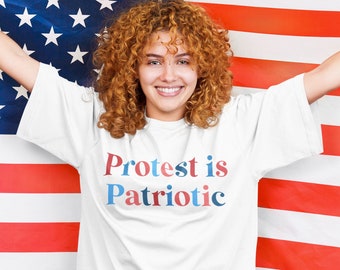 May include: A white t-shirt with the text "Protest is Patriotic" in red, blue, and white lettering. The person wearing the shirt is standing in front of an American flag.