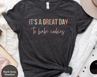 May include: Dark grey heather t-shirt with the text "IT'S A GREAT DAY To bake cookies" in colorful letters. The shirt is knotted at the bottom. A neutral-colored hat and a cup of coffee are in the background.