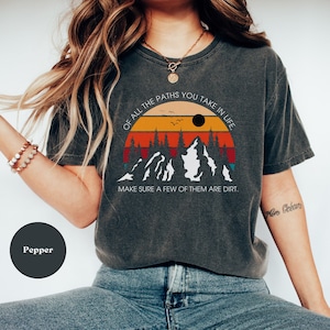 May include: A dark gray t-shirt with a vintage-style graphic of mountains and a sunset. The text on the shirt reads "Of all the paths you take in life, make sure a few of them are dirt."