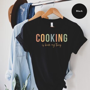 May include: Black t-shirt with the text "COOKING is kinda my thing" in colorful letters.