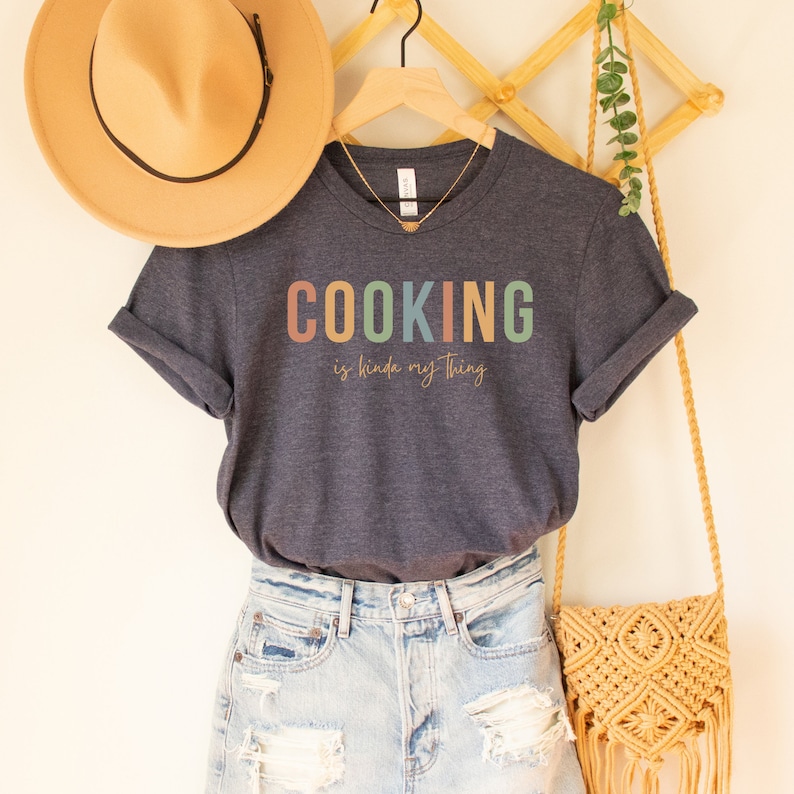 May include: A navy blue t-shirt with the text "COOKING is kinda my thing" in colorful letters. The shirt is hanging on a wooden rack with a straw hat and a woven bag.