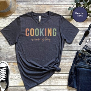 May include: A heather navy blue t-shirt with the text "COOKING is kinda my thing" in rainbow colors. The text is in a retro font style.