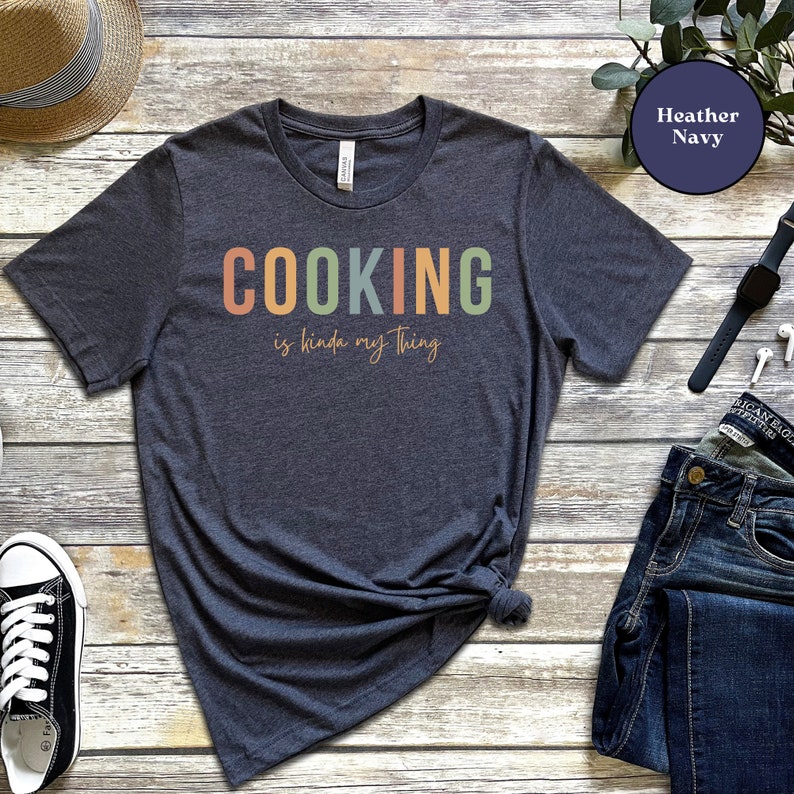 May include: A heather navy blue t-shirt with the text "COOKING is kinda my thing" in rainbow colors. The text is in a retro font style.
