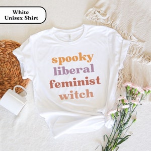 May include: White unisex t-shirt with the text "spooky liberal feminist witch" in orange, purple, and pink.