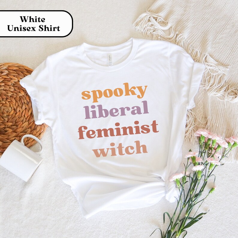 May include: White unisex t-shirt with the text "spooky liberal feminist witch" in orange, purple, and pink.