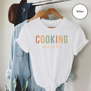 May include: White t-shirt with the text "COOKING is kinda my thing" in rainbow colors.